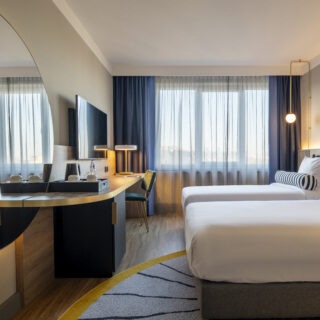 Hotel room with modern decor, featuring a large bed, stylish furniture, neutral color palette, floor-to-ceiling windows with city view, and contemporary artwork on the walls.