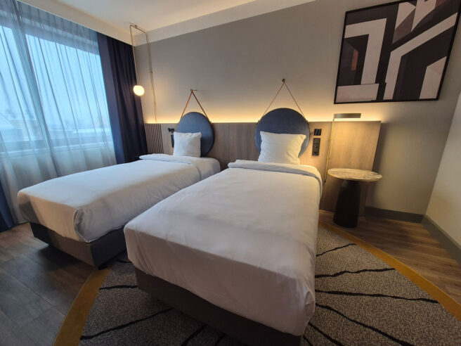A modern hotel room featuring two neatly made twin beds, a desk with a chair, wall-mounted TV, stylish decor, and a window with curtains.